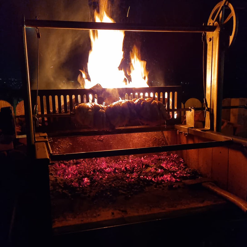 Argentinian grill with glowing embers slowly spinning three chickens over hardwood flames at night